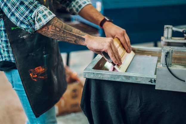 A male screen printing worker with tattooed arms presses ink onto a frame while operating a printing machine.