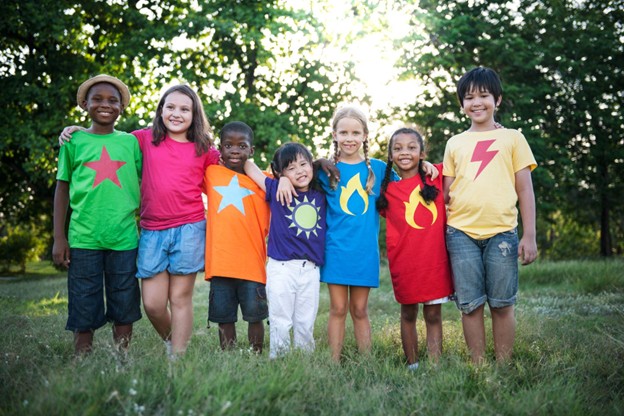 Seven kids wearing vibrantly colored shirts featuring unique T-shirt designs smile with arms around each other.