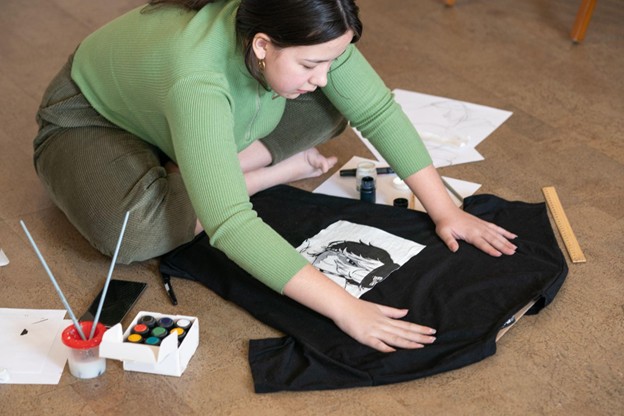 A female designer creates a custom anime-inspired T-shirt design by hand, sketching detailed artwork directly onto the black fabric for a unique clothing piece.