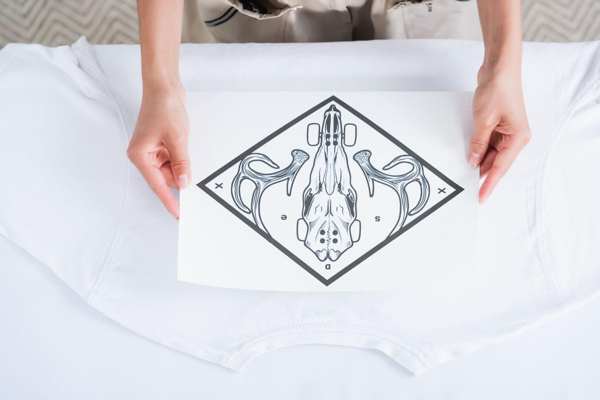 A close-up of a female designer's hands holding a printed design, placed on a blank white shirt, ready for the transfer process.